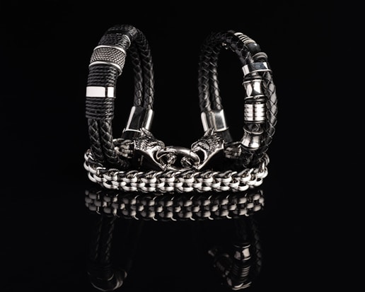 Two intricately designed bracelets composed of black braided leather and shiny metallic components are displayed against a dark reflective surface. One bracelet features a bold chain design with a detailed clasp resembling a wolf’s head.