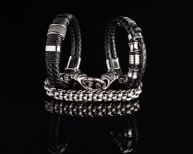 Two intricately designed bracelets composed of black braided leather and shiny metallic components are displayed against a dark reflective surface. One bracelet features a bold chain design with a detailed clasp resembling a wolf’s head.