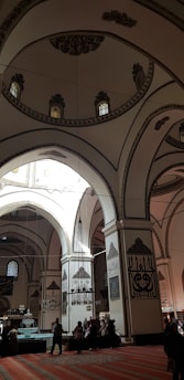 An ornate interior showcases intricate Islamic architectural elements with high arches and decorated walls featuring Arabic calligraphy. Soft light filters through small stained glass windows, illuminating the patterned carpet below. People are visible in silhouette, adding a sense of scale to the spacious and reverent setting.