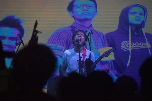 A musician passionately performs on stage with a guitar, surrounded by colorful lights and an audience silhouetted in the foreground. Behind the performer, a large screen displays artistic, posterized images of various individuals.