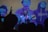 A musician passionately performs on stage with a guitar, surrounded by colorful lights and an audience silhouetted in the foreground. Behind the performer, a large screen displays artistic, posterized images of various individuals.