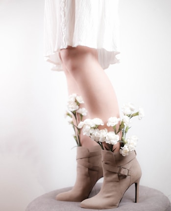 A pair of beige high-heeled boots with white flowers emerging from inside them. The background is softly lit, and part of a white dress is visible. The overall aesthetic is minimalistic and artistic.