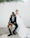 A bride and groom pose together indoors against a plain white background. The groom is seated in a chair, wearing a dark suit with a patterned shirt and holding a bright bouquet of flowers in his hand. The bride stands beside him, dressed in a flowing white gown with her arm around his shoulder. Overhead, green foliage hangs decoratively.
