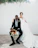 A bride and groom pose together indoors against a plain white background. The groom is seated in a chair, wearing a dark suit with a patterned shirt and holding a bright bouquet of flowers in his hand. The bride stands beside him, dressed in a flowing white gown with her arm around his shoulder. Overhead, green foliage hangs decoratively.