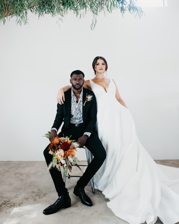 A bride and groom pose together indoors against a plain white background. The groom is seated in a chair, wearing a dark suit with a patterned shirt and holding a bright bouquet of flowers in his hand. The bride stands beside him, dressed in a flowing white gown with her arm around his shoulder. Overhead, green foliage hangs decoratively.