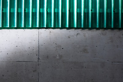 A concrete wall with a green corrugated sheet above it. The wall has a textured surface with visible holes and marks. The corrugated sheet is translucent and contrasts with the wall.