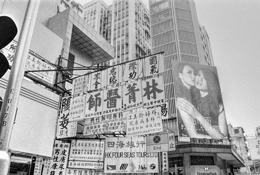 A bustling urban scene with numerous signs and advertisements featuring Chinese characters. Tall buildings with modern architecture dominate the background. One prominent billboard displays an image of two women embracing.