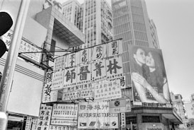 A bustling urban scene with numerous signs and advertisements featuring Chinese characters. Tall buildings with modern architecture dominate the background. One prominent billboard displays an image of two women embracing.