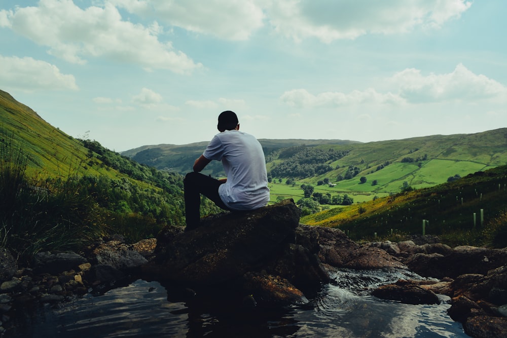 Hombre con camisa blanca sentado en una roca frente al río durante el día