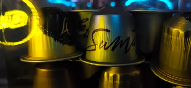 Metallic coffee capsules are stacked closely together with one prominently displaying the word 'Suman'. The capsules have a reflective surface and are illuminated by soft, colored lighting, creating a dynamic interplay of light and shadows.