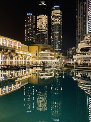 Tall skyscrapers illuminated at night with their lights reflecting in a calm body of water. The buildings, surrounded by modern architecture, include prominent structures that display the name Emaar. The scene is vibrant, with well-lit pathways and lush greenery lining the waterfront.