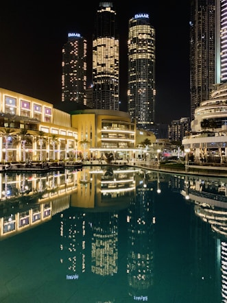 Tall skyscrapers illuminated at night with their lights reflecting in a calm body of water. The buildings, surrounded by modern architecture, include prominent structures that display the name Emaar. The scene is vibrant, with well-lit pathways and lush greenery lining the waterfront.