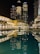 Tall skyscrapers illuminated at night with their lights reflecting in a calm body of water. The buildings, surrounded by modern architecture, include prominent structures that display the name Emaar. The scene is vibrant, with well-lit pathways and lush greenery lining the waterfront.