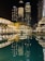 Tall skyscrapers illuminated at night with their lights reflecting in a calm body of water. The buildings, surrounded by modern architecture, include prominent structures that display the name Emaar. The scene is vibrant, with well-lit pathways and lush greenery lining the waterfront.