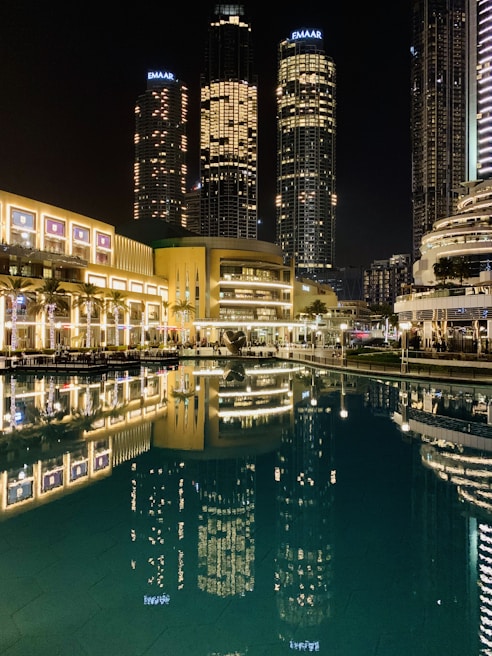 Tall skyscrapers illuminated at night with their lights reflecting in a calm body of water. The buildings, surrounded by modern architecture, include prominent structures that display the name Emaar. The scene is vibrant, with well-lit pathways and lush greenery lining the waterfront.