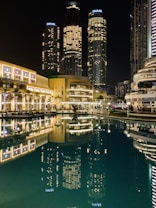 Tall skyscrapers illuminated at night with their lights reflecting in a calm body of water. The buildings, surrounded by modern architecture, include prominent structures that display the name Emaar. The scene is vibrant, with well-lit pathways and lush greenery lining the waterfront.