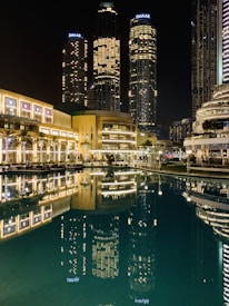 Tall skyscrapers illuminated at night with their lights reflecting in a calm body of water. The buildings, surrounded by modern architecture, include prominent structures that display the name Emaar. The scene is vibrant, with well-lit pathways and lush greenery lining the waterfront.