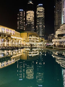 Tall skyscrapers illuminated at night with their lights reflecting in a calm body of water. The buildings, surrounded by modern architecture, include prominent structures that display the name Emaar. The scene is vibrant, with well-lit pathways and lush greenery lining the waterfront.
