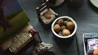 A collection of objects is arranged on a table, including a small container filled with walnuts, a miniature decorative chair, and a textured object resembling coral. There are also various painted and crafted items with intricate designs.