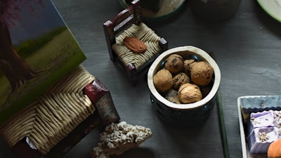 A collection of objects is arranged on a table, including a small container filled with walnuts, a miniature decorative chair, and a textured object resembling coral. There are also various painted and crafted items with intricate designs.