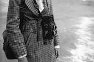 A person wearing a houndstooth patterned coat holds a vintage twin-lens reflex camera hanging from their neck. The image is in black and white, capturing a classic and timeless feel. The background is blurred, focusing on the detailed textures of the coat and the camera.