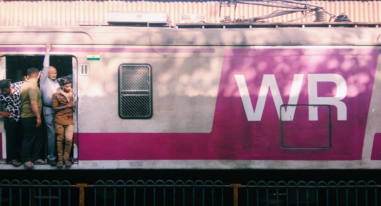 Several people are standing at the open door of a train painted with a large 'WR' logo. The train is light grey with a prominent pink stripe, and there are visible shadows of trees cast on its side. A small Indian flag is displayed next to the door.