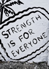 A mural features the phrase 'STRENGTH IS FOR EVERYONE' in bold black letters against a white brick wall. The image includes an artistic depiction of a palm tree and wavy lines surrounding the text.
