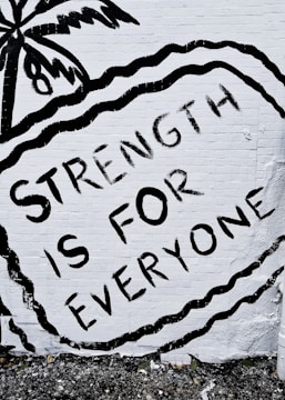 A mural features the phrase 'STRENGTH IS FOR EVERYONE' in bold black letters against a white brick wall. The image includes an artistic depiction of a palm tree and wavy lines surrounding the text.