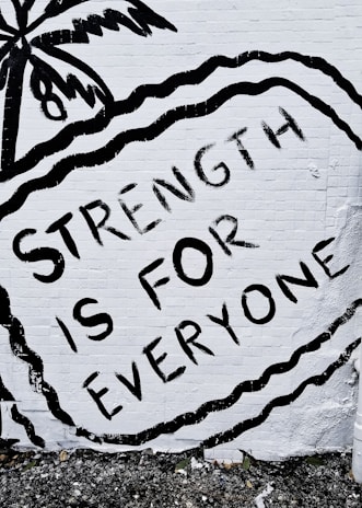 A mural features the phrase 'STRENGTH IS FOR EVERYONE' in bold black letters against a white brick wall. The image includes an artistic depiction of a palm tree and wavy lines surrounding the text.