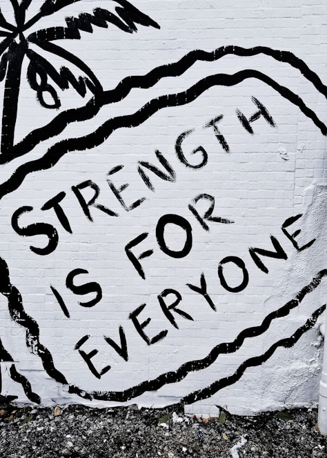 A mural features the phrase 'STRENGTH IS FOR EVERYONE' in bold black letters against a white brick wall. The image includes an artistic depiction of a palm tree and wavy lines surrounding the text.