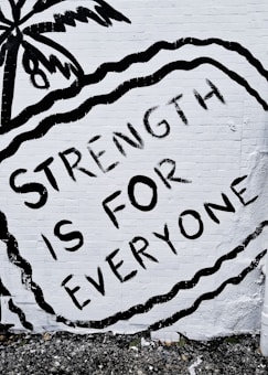 A mural features the phrase 'STRENGTH IS FOR EVERYONE' in bold black letters against a white brick wall. The image includes an artistic depiction of a palm tree and wavy lines surrounding the text.