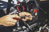 Hands are seen using a screwdriver to work on machinery or a mechanical component. The focus is on the tools and hands, with some wires and connectors visible.