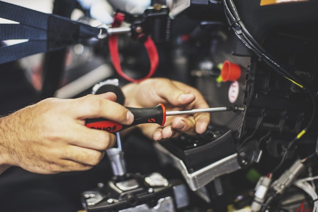 Hands are seen using a screwdriver to work on machinery or a mechanical component. The focus is on the tools and hands, with some wires and connectors visible.