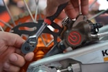 A close-up view of a person using a ratchet wrench to adjust a component on a mechanical device, likely a motorcycle or bicycle brake system. The wrench is being held by a hand, and the caliper has a red logo with visible metal parts and bolts.