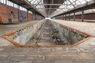 An abandoned industrial facility with a long, narrow pit in the center. The structure features exposed steel beams and broken railroad tracks. Brick walls, adorned with graffiti art, line the interior. The roof is made of large glass panels partially allowing light to enter, casting shadows across the concrete surfaces.