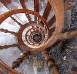 An aerial view of a wooden spiral staircase with worn steps and visible debris. The railings consist of evenly spaced balusters, and the spiral pattern creates a visually striking effect.