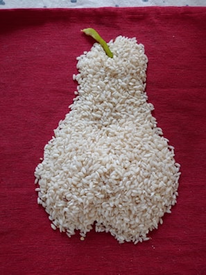 A mound of white rice is arranged in the shape of a pear with a green stem, placed on a textured, deep red fabric background.