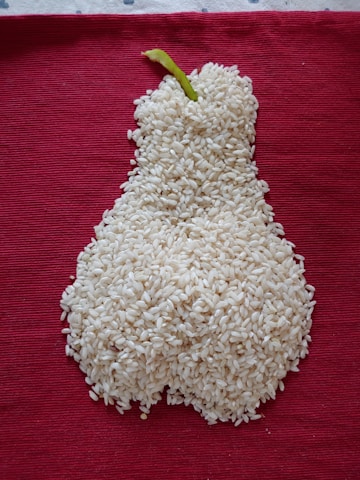 A mound of white rice is arranged in the shape of a pear with a green stem, placed on a textured, deep red fabric background.