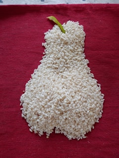 A mound of white rice is arranged in the shape of a pear with a green stem, placed on a textured, deep red fabric background.