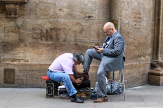 A man in a pink shirt and blue jeans is polishing the shoes of another man seated on a stool, dressed in a gray suit. The man in the suit is using a smartphone, while the shoe polisher is focused on his work. They are situated on a sidewalk against a rustic stone wall.
