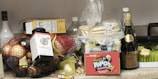 An assortment of groceries and items are placed on a countertop. There are various fruits like apples and avocados in a bowl, a package of tortillas, a box of Hello Panda chocolate snacks, a bottle of supplements, and several bottles of beverages including wine and liqueur. Additionally, a package of kitchen sponges is visible.