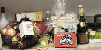 An assortment of groceries and items are placed on a countertop. There are various fruits like apples and avocados in a bowl, a package of tortillas, a box of Hello Panda chocolate snacks, a bottle of supplements, and several bottles of beverages including wine and liqueur. Additionally, a package of kitchen sponges is visible.