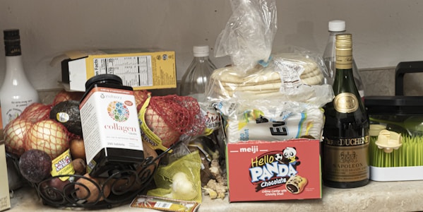 An assortment of groceries and items are placed on a countertop. There are various fruits like apples and avocados in a bowl, a package of tortillas, a box of Hello Panda chocolate snacks, a bottle of supplements, and several bottles of beverages including wine and liqueur. Additionally, a package of kitchen sponges is visible.
