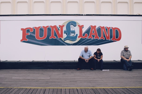 A large white wall with a colorful sign that reads 'FUNLAND' in bold letters. Below the sign, three people are sitting on a wooden bench positioned against the wall. The setting appears to be relaxed, with casual seating.