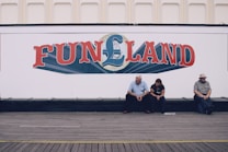 A large white wall with a colorful sign that reads 'FUNLAND' in bold letters. Below the sign, three people are sitting on a wooden bench positioned against the wall. The setting appears to be relaxed, with casual seating.