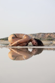 A person with tattoos is in a curled-up position on the ground near a body of water. The serene landscape features a clear reflection in the water, capturing the person's pose with the background of a distant hill. The atmosphere is calm and introspective.