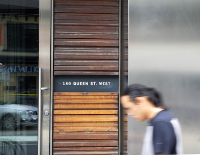 A blurred person walks past a modern building facade, featuring horizontal wooden slats and a sign that reads '180 Queen St. West'. A reflection of a car is visible in the adjacent glass.