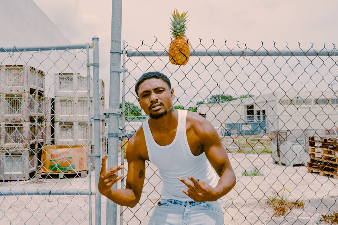 man in white tank top standing beside gray metal fence during daytime