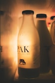 A glass milk bottle with the label PAK is illuminated in a dimly lit environment. Other similar bottles are visible in the background, creating a warm and soft ambiance. The bottle showcases a small cow illustration on the label.