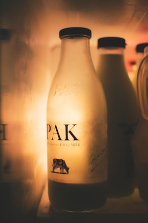 A glass milk bottle with the label PAK is illuminated in a dimly lit environment. Other similar bottles are visible in the background, creating a warm and soft ambiance. The bottle showcases a small cow illustration on the label.